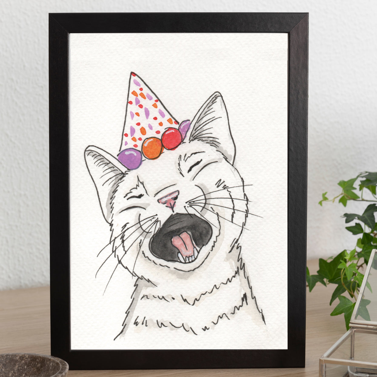 Framed illustration of a cat wearing a colorful party hat on a white wall.
